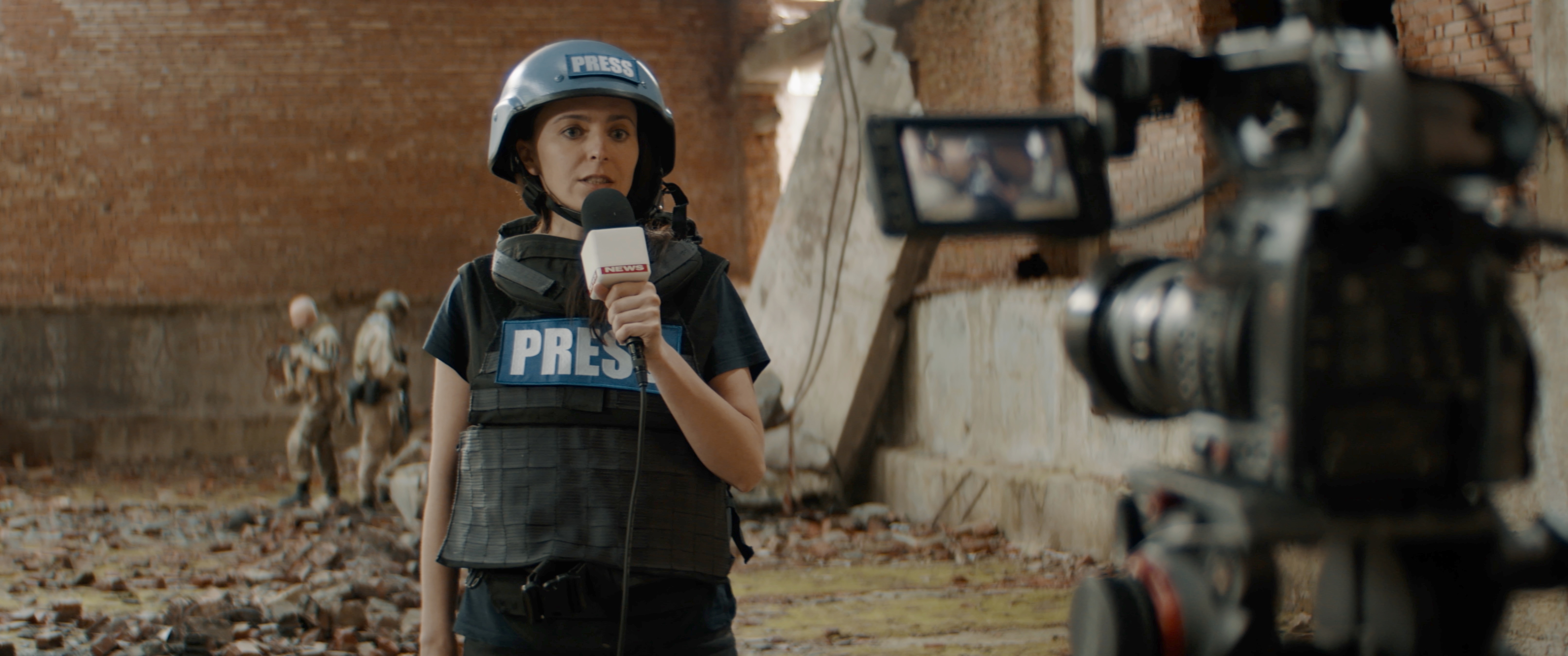 A reporter wearing a PRESS helmet and vest speaking to camera in a damaged building.